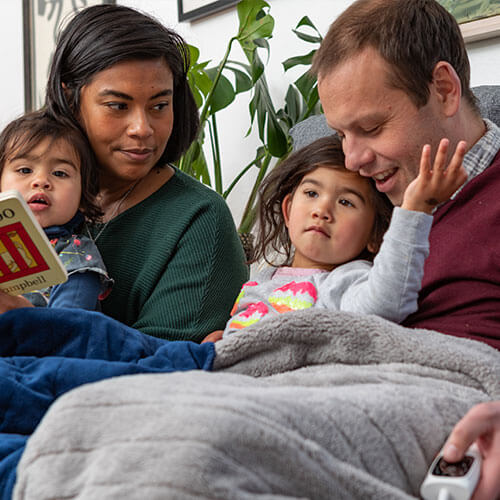 A young mother and father cuddling on the couch with their two toddlers. They're under a heated blanket and reading a storybook together.