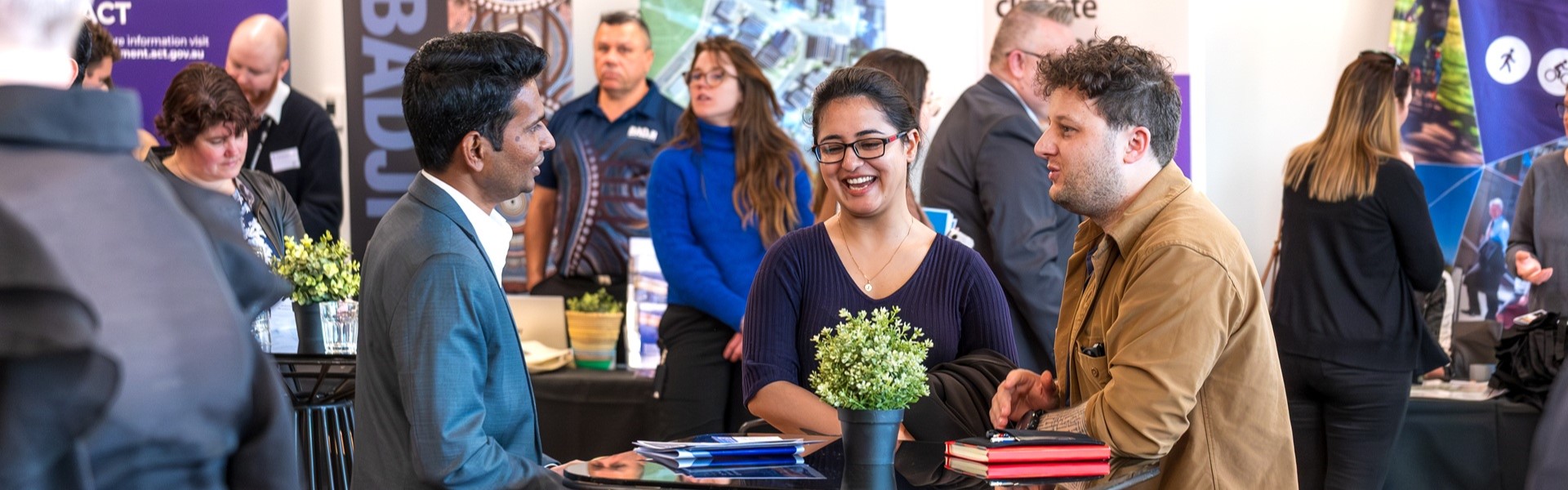 People talking at a business networking event around tables with banners, and information materials in the background.