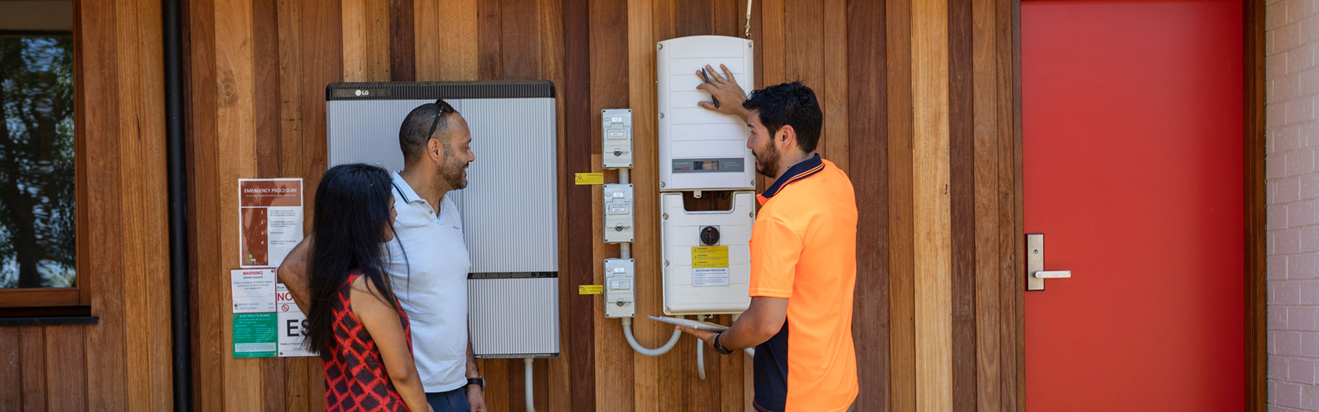 An installer in an orange hi-vis vest explaining a household storage battery on the side of a home to a young couple.