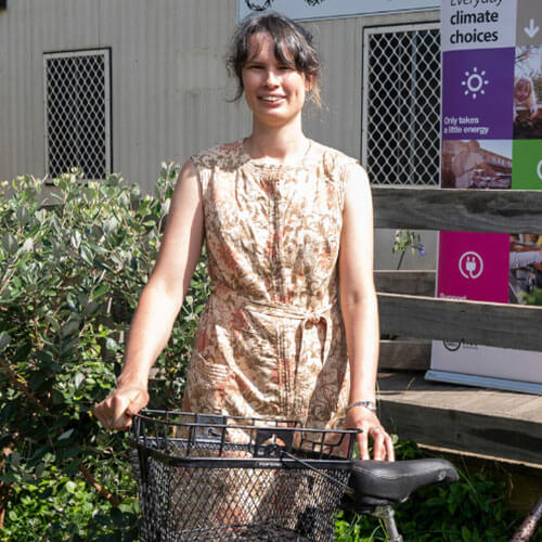A photo of Canberra Environment Centre acting Director, Zoe, standing outside the ReCyclery building holding the handlebars of a bike. An Everyday Climate Choices pull up banner is in the background.