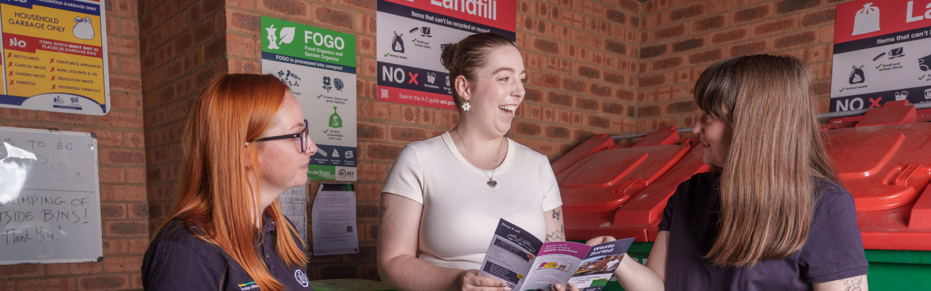 Three people are standing inside a brick bin enclosure, holding and looking at waste education brochures. Behind them are large red-lidded landfill bins and wall posters with waste sorting information, including signs for landfill and Food Organics and Garden Organics (FOGO). A whiteboard with handwritten notes is visible on the left side of the image.