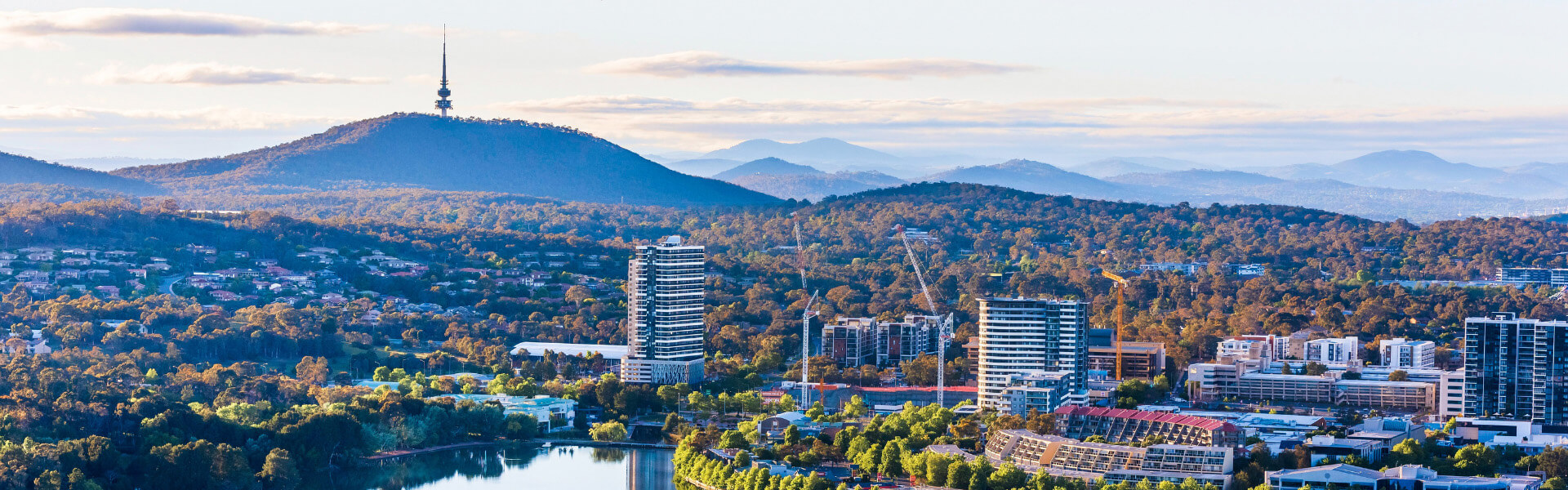 Panoramic view of Canberra looking over Belconnen. The town centre and Lake Ginninderra is in the foreground. Black Mountain and Telstra Tower are prominent in the background. In the far distance are mountain ranges. It is early morning, the sky has become blue and small clouds hang over the mountains in the background.    