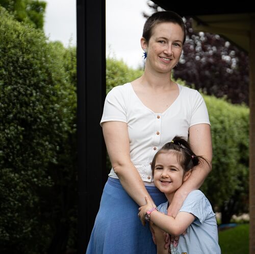 Girl standing in front of mother with arms around her in front of a verandah post and hedge.