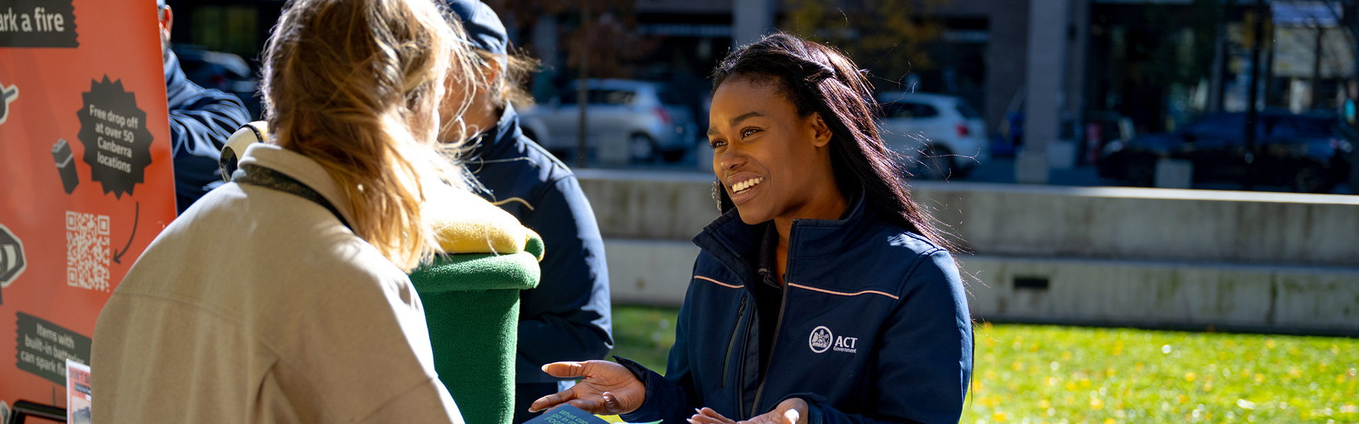 A person wearing a blue jacket with the ACT logo is speaking to another person who is holding a green object. The background shows an outdoor setting with grass, trees, and parked cars. 