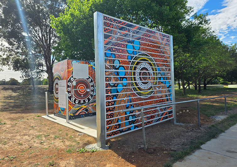 A small utility structure surrounded by metal railings and decorated with vibrant Indigenous-style artwork featuring concentric circles, dot patterns, and bright colours such as orange, blue, black, and white. A tall metal screen beside the structure is also covered in the same artwork. The structure sits on a concrete pad in a park-like setting with trees, grass, and a partly cloudy sky in the background.
